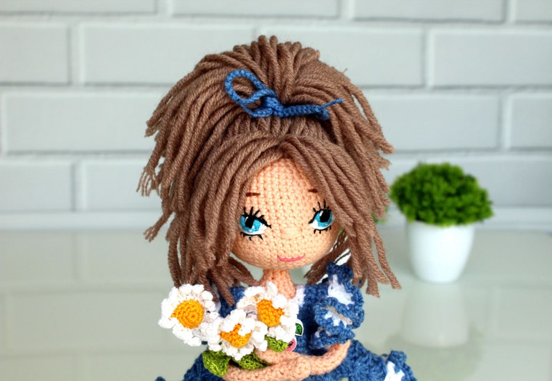 Peut inclure: Une poup&eacute;e en crochet avec des cheveux bruns, des yeux bleus et un n&oelig;ud bleu. La poup&eacute;e porte une robe bleue avec des accents blancs et tient un bouquet de marguerites blanches.