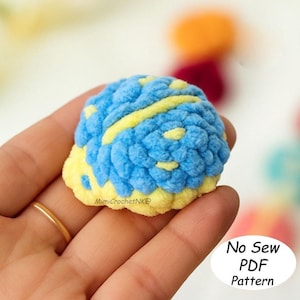 May include: A small, handmade crochet item in shades of blue and yellow. The textured, round object has a fluffy appearance, with a prominent yellow stripe. The text "No Sew PDF Pattern" is visible in a white oval. The item is held in a hand.