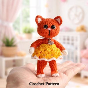 May include: A crocheted orange cat doll with white detailing on the ears and paws, wearing a yellow skirt. The doll has black eyes and a small pendant. The words "Crochet Pattern" are at the bottom.