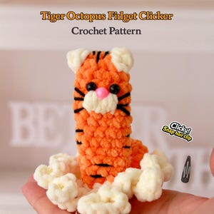 May include: A crocheted tiger octopus fidget clicker in orange with black stripes, a pink nose, and black eyes. The text "Tiger Octopus Fidget Clicker Crochet Pattern" is at the top. A snap hair clip is also visible.