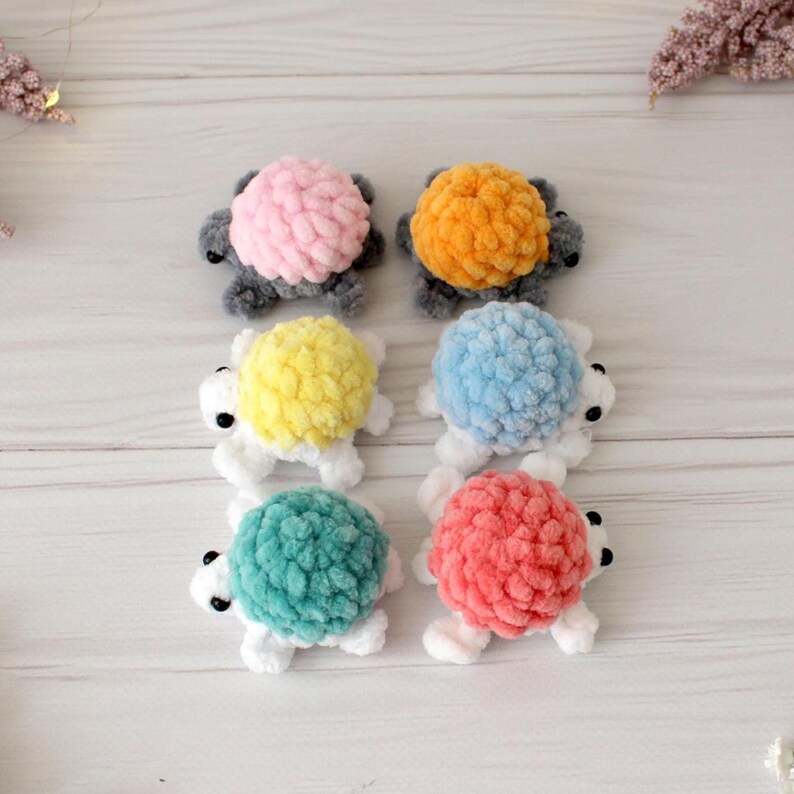 May include: Six handmade crochet turtle toys in various colors: pink, orange, yellow, blue, teal, and red. Each turtle has a textured shell and white limbs. The turtles are arranged on a light wooden surface.
