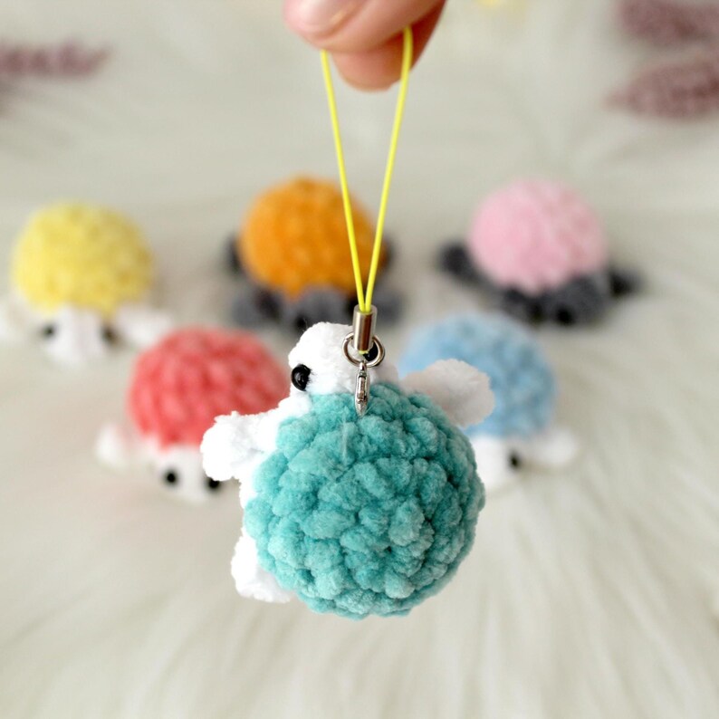 May include: A handmade, plush turtle charm in turquoise, white, and black, held by a yellow cord. Other colorful turtles in yellow, orange, pink, red, and blue are in the background. The turtles have a soft, fuzzy texture.