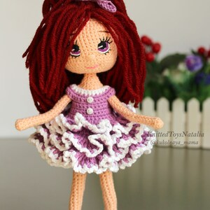 Peut inclure: Une poup&eacute;e en crochet aux longs cheveux roux, v&ecirc;tue d'une robe violette &agrave; volants blancs et de chaussures violettes. La poup&eacute;e a une expression douce avec de grands yeux et un n&oelig;ud rose dans les cheveux. Knitted Toys Natalia @kukolnaya_mama