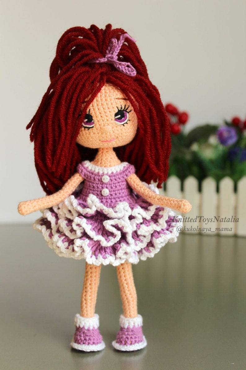 Peut inclure: Une poup&eacute;e en crochet aux longs cheveux roux, v&ecirc;tue d'une robe violette &agrave; volants blancs et de chaussures violettes. La poup&eacute;e a une expression douce avec de grands yeux et un n&oelig;ud rose dans les cheveux. Knitted Toys Natalia @kukolnaya_mama