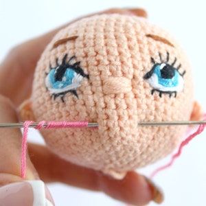 Peut inclure: Gros plan sur une t&ecirc;te de poup&eacute;e en crochet avec des yeux bleus, des sourcils bruns et une bouche rose en cours de couture avec une aiguille et du fil.