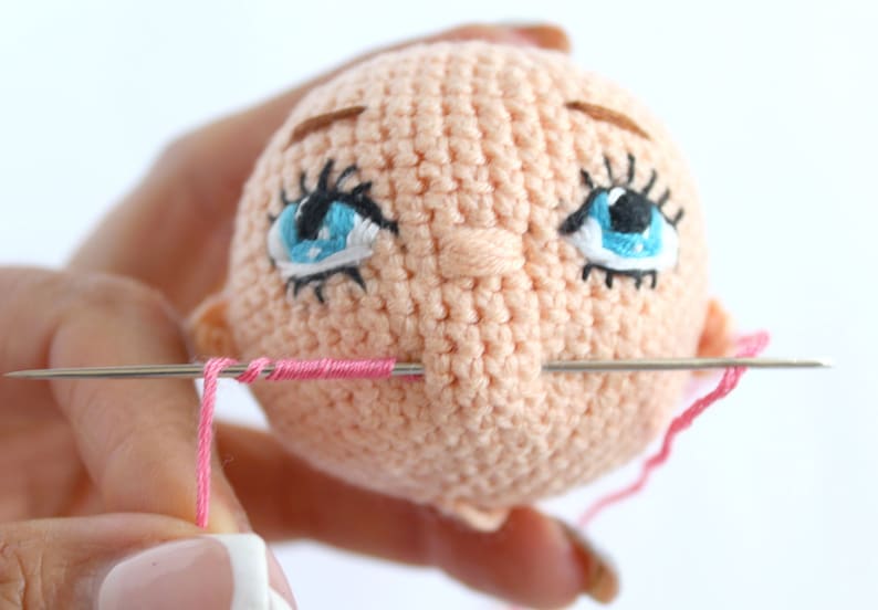 Peut inclure: Gros plan sur une t&ecirc;te de poup&eacute;e en crochet avec des yeux bleus, des sourcils bruns et une bouche rose en cours de couture avec une aiguille et du fil.