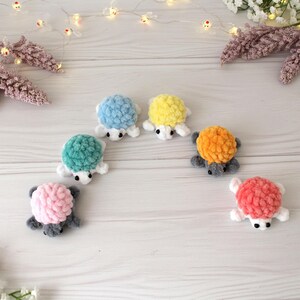 May include: A collection of six handmade crochet turtle toys in various colors: blue, yellow, green, pink, orange, and coral. Each turtle has a textured shell, white flippers, and a gray body. The toys are arranged on a light wooden surface.