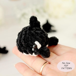 May include: A handmade, crocheted orca whale in black and white. The small plush toy has a rounded body, a dorsal fin, and white markings. The image includes a small, light pink oval with the text "No-Sew PDF PATTERN".