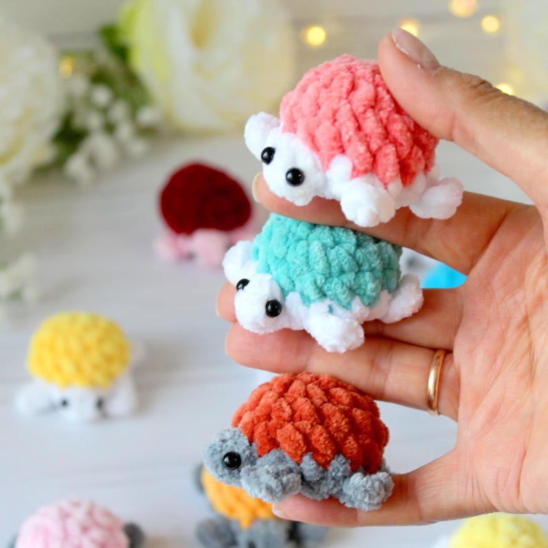 May include: Three small, handmade crochet turtles in shades of pink, teal, and orange are stacked in a hand. Each turtle has a textured shell, white limbs, and black eyes. Other turtles in yellow and pink are visible in the background.