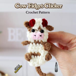 May include: A handmade crochet cow fidget clicker. The cow has a white body with brown spots, brown legs, and red ears. The text "Cow Fidget Clicker Crochet Pattern" is displayed above the cow. A snap hair clip is next to the cow.