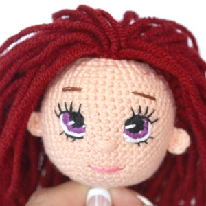 Peut inclure: Gros plan sur la t&ecirc;te d'une poup&eacute;e en crochet avec des cheveux en laine rouge, une peau rose et des yeux violets brod&eacute;s.
