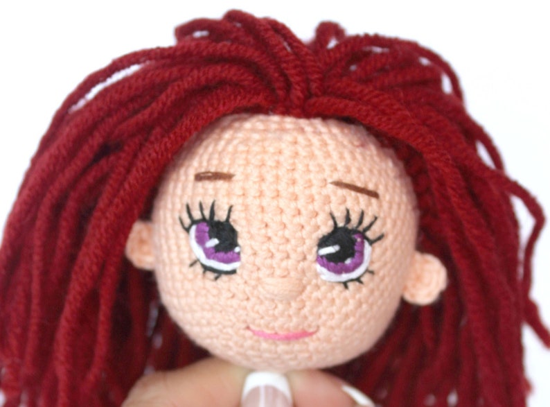 Peut inclure: Gros plan sur la t&ecirc;te d'une poup&eacute;e en crochet avec des cheveux en laine rouge, une peau rose et des yeux violets brod&eacute;s.