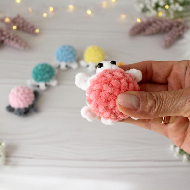 May include: A handmade, plush turtle in coral pink, held in a hand. The turtle has white flippers and head, with black eyes. Other turtles in blue, yellow, green, and pink are in the background, suggesting a collection of soft toys.