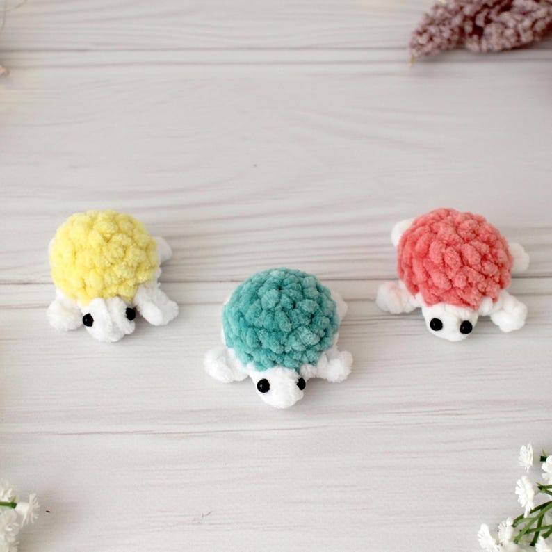 May include: Three handmade crochet turtle figurines in yellow, teal, and coral. Each turtle has a textured shell, white flippers, and black eyes. The turtles are displayed on a light-colored wooden surface.