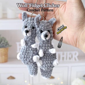 May include: Two gray crochet wolf keychains with white accents, black eyes, and silver keyrings. The text "Wolf Fidget Clicker Crochet Pattern" is at the top. A black snap hair clip is visible. The keychains are held in a hand.