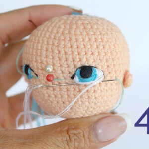 Peut inclure: Gros plan sur une t&ecirc;te de poup&eacute;e en crochet avec des yeux bleus, un nez rose et une perle blanche. La t&ecirc;te de la poup&eacute;e est cousue avec du fil blanc. Le num&eacute;ro 41 est visible dans le coin inf&eacute;rieur droit de l'image.