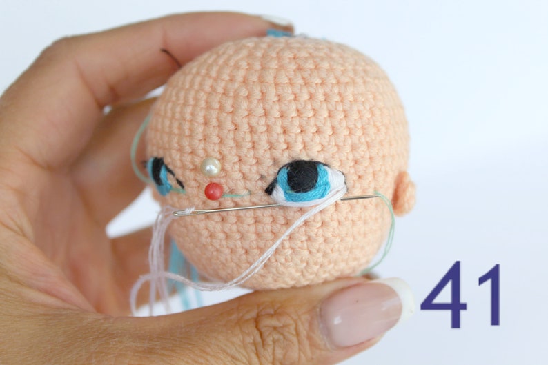 Peut inclure: Gros plan sur une t&ecirc;te de poup&eacute;e en crochet avec des yeux bleus, un nez rose et une perle blanche. La t&ecirc;te de la poup&eacute;e est cousue avec du fil blanc. Le num&eacute;ro 41 est visible dans le coin inf&eacute;rieur droit de l'image.