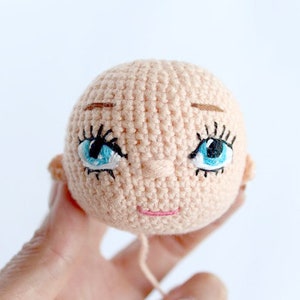 Peut inclure: Une t&ecirc;te de poup&eacute;e en crochet avec des yeux bleus, des sourcils bruns et un sourire rose.