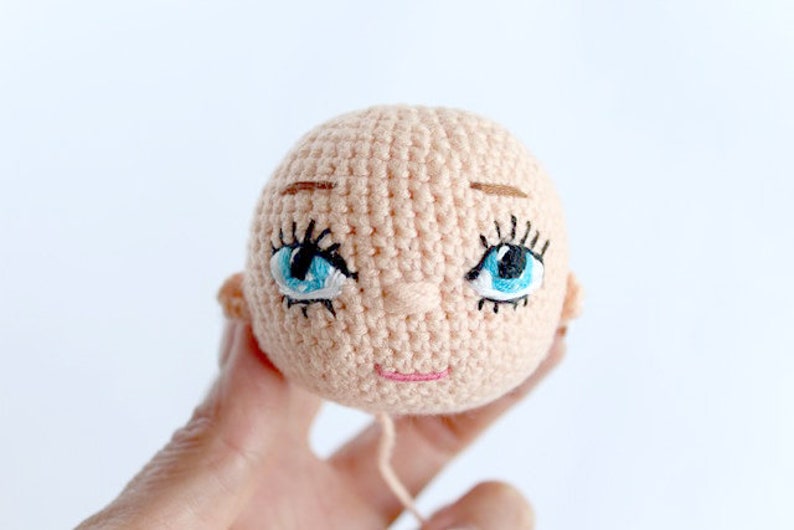 Peut inclure: Une t&ecirc;te de poup&eacute;e en crochet avec des yeux bleus, des sourcils bruns et un sourire rose.