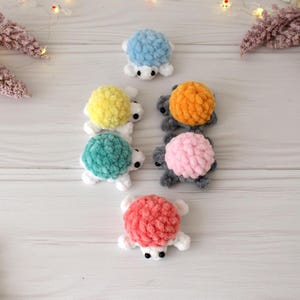 May include: A collection of handmade crochet turtles in various colors, including blue, yellow, teal, orange, pink, and coral. Each turtle has a textured, fluffy shell and white limbs and head. The turtles are arranged on a light wooden surface.