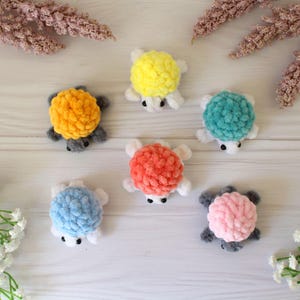 May include: A collection of handmade crochet turtle toys in various colors, including yellow, orange, teal, red, blue, and pink. Each turtle has a textured shell, white flippers, and small black eyes. The toys are arranged on a light-colored surface.