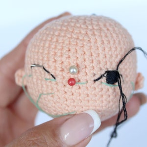 Peut inclure: Gros plan sur une t&ecirc;te de poup&eacute;e en crochet avec du fil noir et vert pour les yeux et une perle blanche pour le nez. La t&ecirc;te de la poup&eacute;e est rose et a une &eacute;pingle rouge au centre du visage.