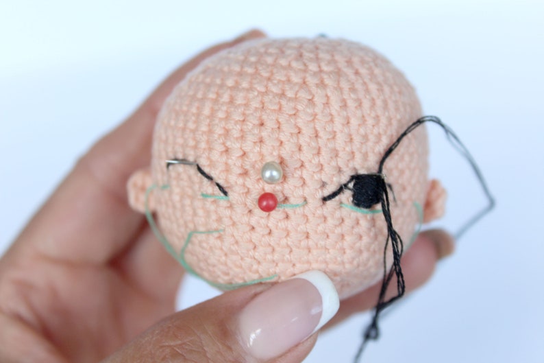 Peut inclure: Gros plan sur une t&ecirc;te de poup&eacute;e en crochet avec du fil noir et vert pour les yeux et une perle blanche pour le nez. La t&ecirc;te de la poup&eacute;e est rose et a une &eacute;pingle rouge au centre du visage.