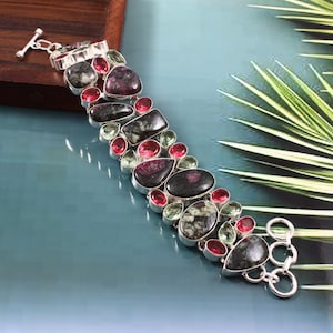 以下が含まれることがあります： トグル留め具が付いたシルバーブレスレットは、緑、ピンク、黒のファセットカットされた宝石が並んでいます。ブレスレットはスターリングシルバー製で、長さは約18cmです。