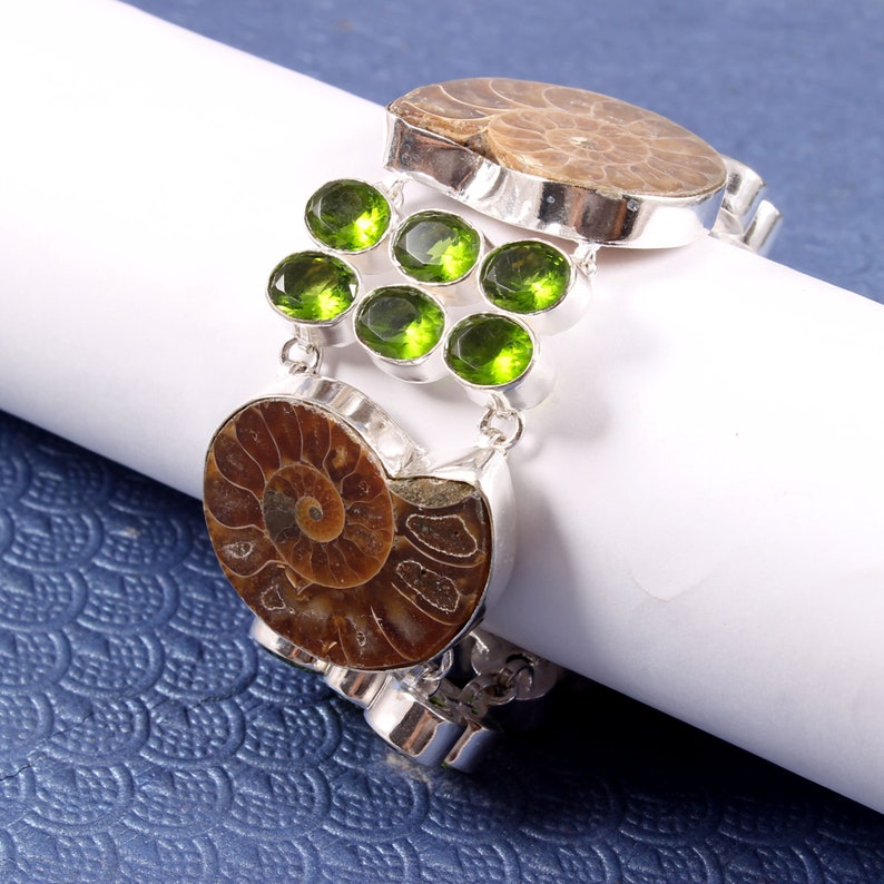 May include: A silver bracelet with two brown ammonite fossils and green gemstones. The bracelet is on a white surface.