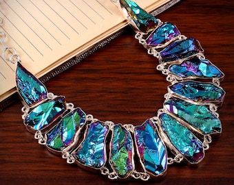 Puede incluir: Un collar de plata con un gran colgante de cristal iridiscente azul, verde y morado. El colgante está formado por varias piezas de cristal que están conectadas entre sí.