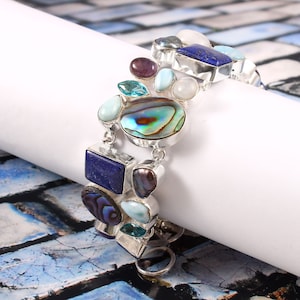 May include: A silver bracelet with a variety of colorful gemstones, including blue lapis lazuli, abalone shell, turquoise, and amethyst. The bracelet has a unique, asymmetrical design.