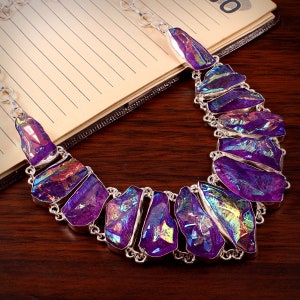 Puede incluir: Un collar de plata con un gran colgante de cristal morado iridiscente. El colgante está hecho de varias piezas de cristal que están conectadas entre sí. El collar está sobre una superficie de madera.