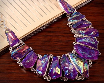 Puede incluir: Un collar de plata con un gran colgante de cristal morado iridiscente. El colgante está hecho de varias piezas de cristal que están conectadas entre sí. El collar está sobre una superficie de madera.