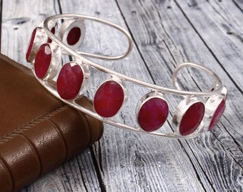 May include: A silver bangle bracelet with a row of oval-shaped red gemstones. The bracelet is open and has a simple design.