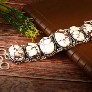 Op de afbeelding: Een zilveren armband met ovale witte en bruine stenen. De stenen zijn gezet in een decoratief zilveren frame. De armband wordt getoond op een houten oppervlak naast een bruin leren notitieboek.