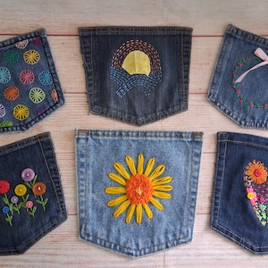May include: Six denim pockets arranged in two rows. The top row features a pocket with colorful embroidered circles, a pocket with a yellow sun and blue stitching, and a pocket with a floral wreath and pink ribbon. The bottom row shows a pocket with button flowers, a large yellow sunflower, and a heart filled with colorful embroidery and buttons.