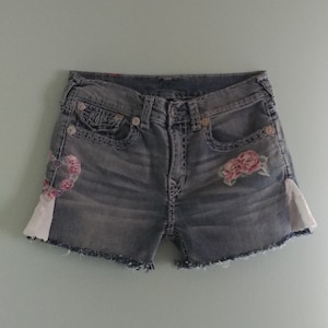 May include: A pair of denim shorts with a frayed hem and floral embroidery. The shorts have a light wash and are decorated with pink roses on the front.