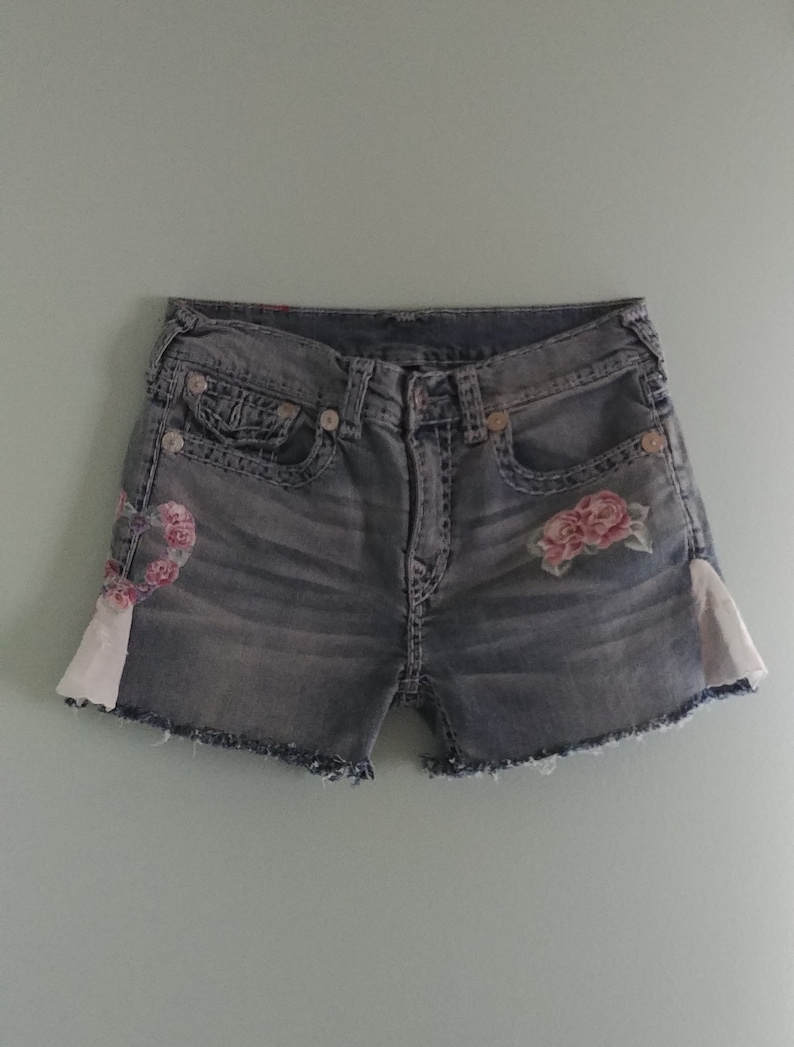 May include: A pair of denim shorts with a frayed hem and floral embroidery. The shorts have a light wash and are decorated with pink roses on the front.