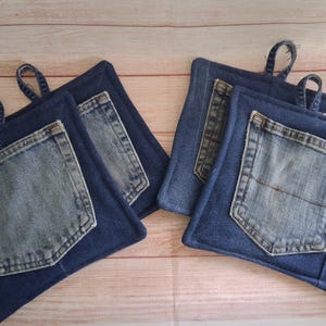 May include: Four square pot holders made from dark blue denim fabric. Each pot holder features a lighter blue denim pocket stitched onto the front and a small denim loop at the top corner for hanging. The edges are finished with blue fabric binding.