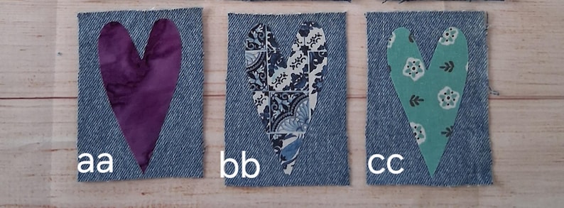 May include: Three denim fabric squares with heart cutouts. The hearts are filled with different patterned fabrics: a purple batik, a blue and white floral, and a teal floral. Each square has a white letter designation.