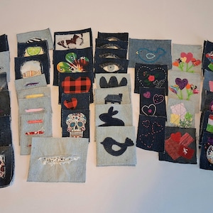 May include: A collection of denim patches with various embroidered and appliqued designs. The patches feature images such as stars, hearts, flowers, a skull, and a bird. The patches are in shades of blue and gray.