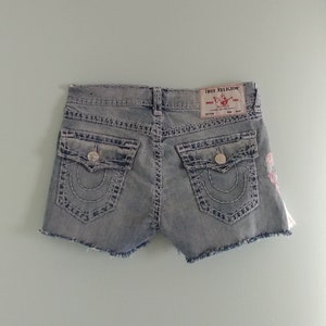 May include: Light blue denim shorts with a frayed hem and embroidered flowers. The shorts have a back pocket with the True Religion logo and a button closure.