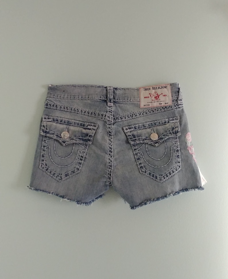May include: Light blue denim shorts with a frayed hem and embroidered flowers. The shorts have a back pocket with the True Religion logo and a button closure.