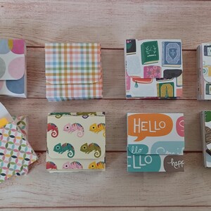 May include: An assortment of small, decorative paper boxes with various patterns, including polka dots, plaid, and chameleons. One box is open, revealing a yellow object. The boxes feature colorful designs and the word "HELLO".