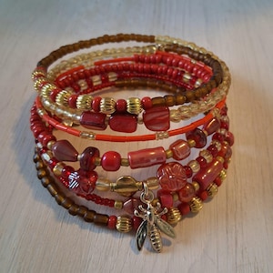 Peut inclure: Un bracelet multi-tours composé de perles dans des tons rouges, marron et dorés. Le design intègre des perles rondes lisses, des perles cylindriques et des éléments en verre facetté. Une breloque abeille argentée est suspendue au centre.