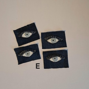 Puede incluir: Cuatro parches de mezclilla, cada uno con un dise&ntilde;o de ojo bordado. Los ojos tienen un iris blanco con una pupila negra, sobre un fondo de mezclilla azul oscuro. La letra "E" es visible.