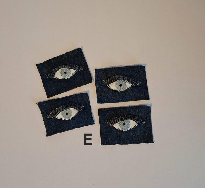 Puede incluir: Cuatro parches de mezclilla, cada uno con un dise&ntilde;o de ojo bordado. Los ojos tienen un iris blanco con una pupila negra, sobre un fondo de mezclilla azul oscuro. La letra "E" es visible.