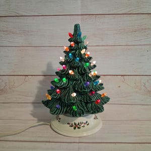 May include: A dark green ceramic Christmas tree lamp with a textured, layered branch design. The tree is decorated with small, multi-coloured glowing plastic bulbs in red, blue, orange, and clear. It sits on a round, cream-coloured base with holly.