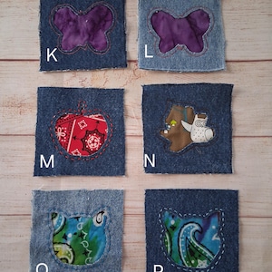 May include: Six denim fabric squares with various appliqued designs. The designs include a purple butterfly, a red apple, a brown bear with an owl, and two cat faces with paisley patterns. Each square is outlined with white stitching.