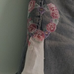 May include: A pair of denim shorts with a floral heart design on the front. The heart is made of pink, purple, and green flowers. The shorts have a frayed hem.
